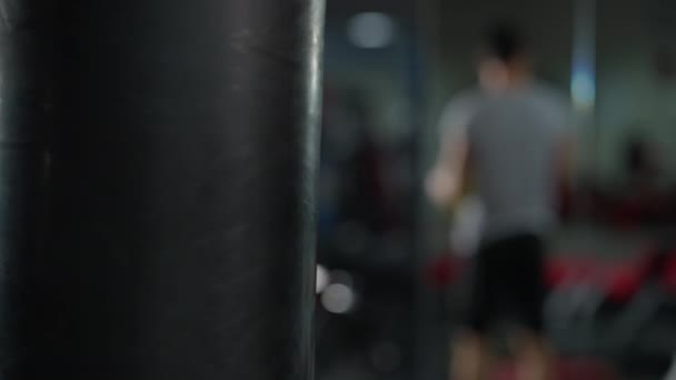 Bokszzsák lóg a tornateremben homályos férfi emel súlyzókat a háttérben. Bezárt boxberendezés. A harcművészetek és az egészséges életmód fogalma. — Stock videók