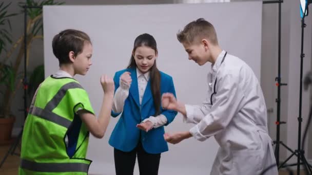 Three cheerful carefree teenage boys and girl playing rock paper scissors smiling having fun. Positive joyful Caucasian teenagers in professions uniforms enjoying leisure together indoors. Lifestyle. — Stock Video