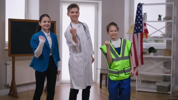 Positivo sorridente adolescente meninos e menina gesticulando vitória sorrindo olhando para a câmera em pé dentro de casa. Três adolescentes caucasianos escolhendo profissões posando em médico construtor e fotógrafo uniforme. — Vídeo de Stock