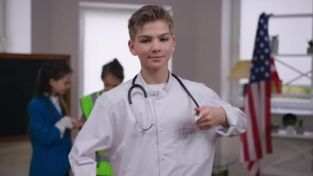 Menino feliz mostrando estetoscópio gesticulando polegar para cima olhando para a câmera sorrindo com amigos borrados no fundo. Retrato de adolescente caucasiano positivo confiante posando escolhendo trabalho de sonho. — Vídeo de Stock