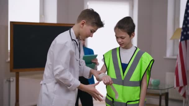 Anak remaja kaukasia dengan gaun dokter tangan bandaging teman dalam seragam pembangun. Remaja yang percaya diri positif memilih profesi sebagai praktisi. Konsep gaya hidup dan pengambilan keputusan. — Stok Video