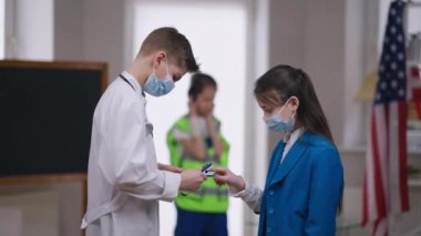 Doktor cüppeli genç çocuk ve Covid-19 yüz maskesi kız konuşmasının oksijen doygunluğunu ölçmek için nabız oksimetresi kullanıyor. Kendine güvenen Kafkas pozitif genç salgın hastalıklar üzerine tıp mesleğini seçiyor..