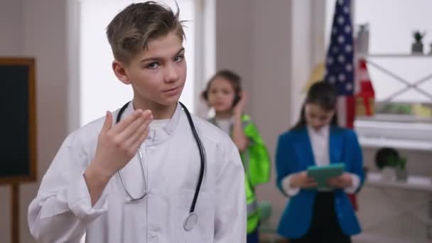 Serious teenage boy putting on coronavirus face mask gesturing thumb up looking at camera. Portrait of confident Caucasian teenager on medical gown aspiring safety protection on Covid-19 pandemic. — Stock Video