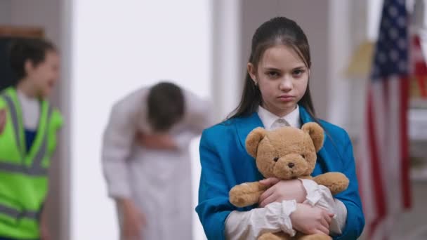 Szomorú zaklatott lány ölelgeti Teddy macit, miközben a fiúk a háttérre mutogatnak. Depressziós, fehér, gyönyörű tinédzser portréja. Zaklatás és szomorúság fogalma. — Stock videók