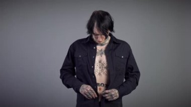 Siyah gömlekli dövmeli genç adamın portresi kendine güvenen yüz ifadesiyle kameraya bakıyor. Kafkasyalı esmer adam gri arka planda poz veriyor..