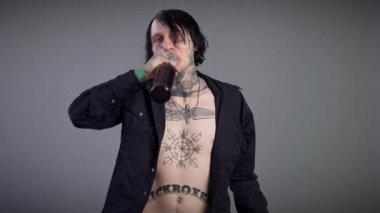 Düğmeleri iliklenmemiş genç adam kameraya bakarak şişeden bira içiyor. Gri arka planda alkol şişesi olan kendine güvenen beyaz adamın portresi..