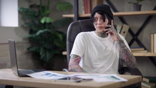 Allegro uomo tatuato in maglietta bianca e cappello nero che parla al telefono seduto in ufficio in isolamento coronavirus. Ritratto di felice allegro freelance caucasico che discute il progetto sorridendo. — Video Stock
