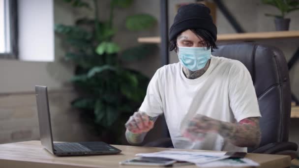 Mladý tetovaný muž v klobouku a Covid-19 maska psaní na klávesnici notebooku analyzující papírování zpráv on-line na dálkové práci. Portrét koncentrovaného profesionálního nezávislého pracovníka uvnitř. — Stock video