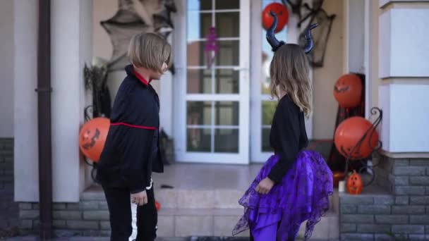 Живая камера следит за веселыми мальчишками и девчонками в костюмах Хэллоуина, бегущими в украшенный дом на праздник. Счастливого беззаботного кавказского брата и сестру, наслаждающихся праздником. — стоковое видео