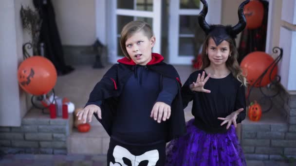 Anak laki-laki dan perempuan Kaukasia yang ceria dengan kostum Halloween kerangka vampir dan setan yang menyeringai membuat wajah-wajah melihat kamera berdiri di luar ruangan. Teman hiperaktif bersenang-senang dengan pose liburan. Gerakan lambat — Stok Video