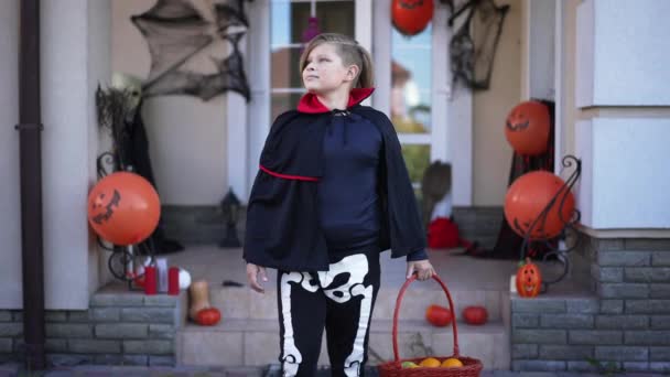 Happy Caucasian little boy in Halloween vampire skeleton costume walking in slow motion with basket leaving. Smiling cute child strolling on front yard from decorated house. Happiness and childhood. — Stock Video
