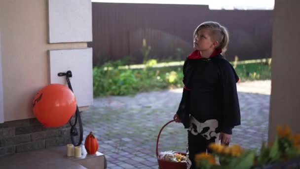 Симпатичный мальчик с корзиной конфет или конфет входит в украшенное крыльцо на переднем дворе, оглядываясь вокруг уходя. Портрет счастливого кавказского ребенка, идущего по соседству на Хэллоуин в костюме вампира. — стоковое видео