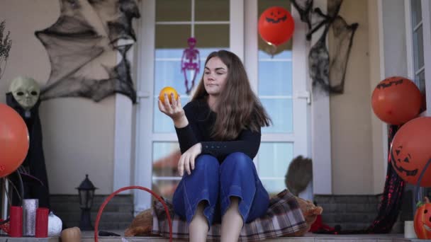 Retrato de la joven feliz hermosa mujer caucásica sentada en el porche del patio trasero en Halloween oliendo naranja sonriente. Relajada dama milenaria disfrutando de la celebración de las vacaciones en la casa decorada. Movimiento lento. — Vídeos de Stock