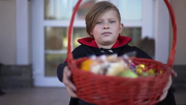 Front view portrait of cute boy posing with Trick or Treat basket looking at camera sitting outdoors. Happy Caucasian kid showing sweets candies fruits bragging Halloween treats. Slow motion. — Stock Video