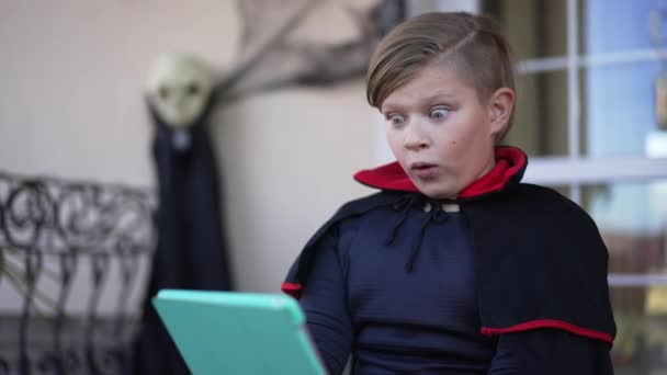 Caucasian little boy making scared face watching horror on tablet sitting outdoors on porch. Portrait of cute kid celebrating Halloween in vampire costume enjoying leisure. Slow motion. — Stock Video