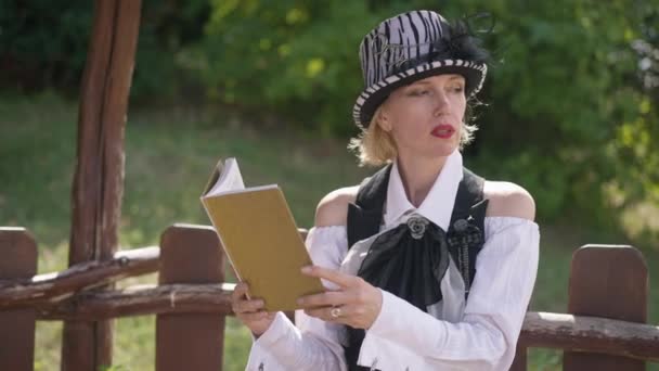 Mooie volwassen vrouw in steampunk kostuum en hoed denken buiten zitten op zonnige dag en het lezen van boek. Portret van slimme zelfverzekerde blanke dame die geniet van hobby. Levensstijl en eenzaamheid. — Stockvideo