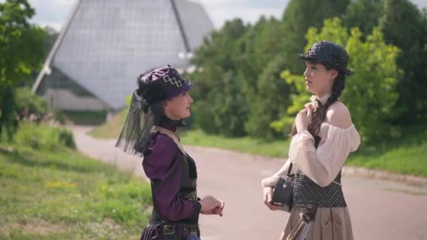Side view two stylish women in steampunk costumes standing outdoors talking smiling. Positive Caucasian ladies cosplaying enjoying sunny day on Halloween. Style and lifestyle. — Stock Video