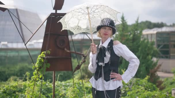 Donna sicura di sé a tiro medio in abiti steampunk in bianco e nero in piedi con ombrellone all'aperto. Ritratto di sottile attraente signora caucasica cosplaying in futuristico stile post-apocalittico. — Video Stock