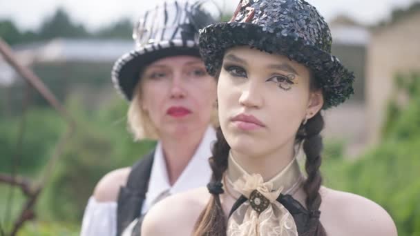 Närbild allvarligt ansikte ung smal vacker vit kvinna i steampunk outfit poserar utomhus med suddig vuxen dam i bakgrunden. Dotter och mor på halloween. — Stockvideo