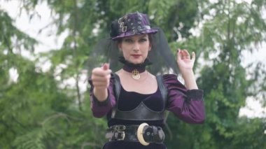 Canlı kameraya yakından bakarken orta boy, ince, beyaz bir cadının görüntüsü. Steampunk kostümlü güzel, zayıf bir kadının cadılar bayramında açık havada poz vermeye davet edildiği portresi. Yavaş çekim.