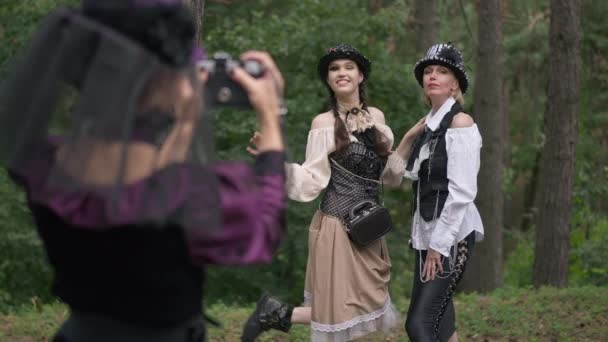 Mujeres delgadas posando para un amigo tomando fotos en la cámara en el bosque al aire libre. Señoras caucásicas seguras positivas en traje steampunk de Halloween disfrutando de la celebración navideña en los bosques. Movimiento lento. — Vídeo de stock