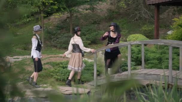 Live camera follows three smiling confident women in witches Halloween costumes passing wooden bridge over stream in woods. Wide shot happy Caucasian ladies in steampunk outfit on holiday. — Stock Video
