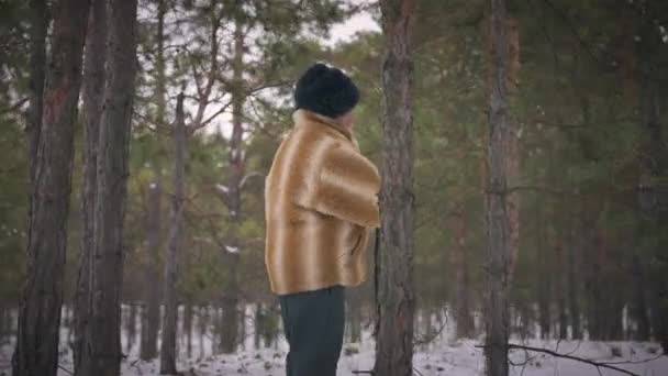 Oldalnézet idősebb férfi szőrmebundában sétál havas téli erdőben lassított felvételen. Egy öreg férfi felfedező sétál a fenyőerdőben a szabadban. Természet és életmód fogalma. — Stock videók