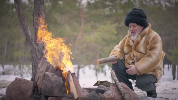 Viejo indígena sonriente sentado en el bosque de invierno al fuego con leña. Amplio retrato de un hombre mongol mayor y seguro al aire libre en un día frío. Movimiento lento. — Vídeos de Stock