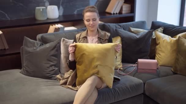 Portrait of smiling happy Caucasian slim beautiful woman admiring and hugging decorative mustard cushion sitting on couch in furniture showroom. Attractive female buyer making choice in shop. — Stock Video