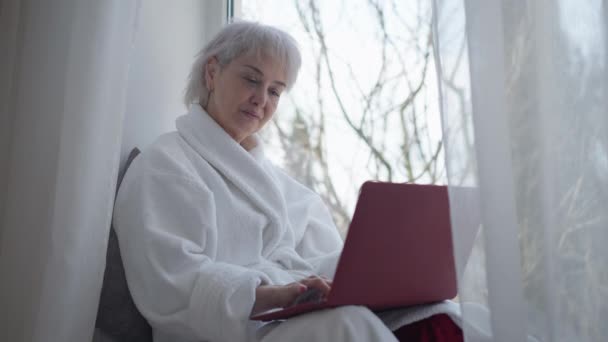 Koncentrált, érett nő ül az ablakpárkányon, gépel a laptop billentyűzetén, szörfözik az interneten. Portré intelligens kaukázusi szabadúszóról, aki otthon online dolgozik. Távmunka-koncepció. — Stock videók