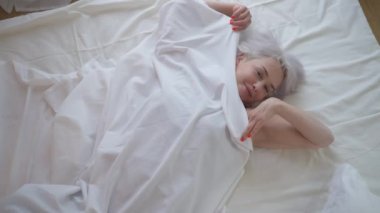 Üst manzara, yatakta uzanmış kameraya bakarak kendine güvenen olgun bir kadına yakınlaşır. Güzel, ince, beyaz bir kadın sabah yatak odasında battaniyeyle kapanış pozu veriyor. Ev hayatı kavramı.