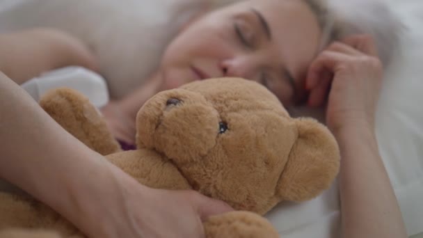 Detailní záběr medvídek s rozmazané dospělé ženy spí v posteli v pozadí. Klidná kavkazská štíhlá dáma odpočívající o víkendu ráno doma v ložnici s hračkou. — Stock video