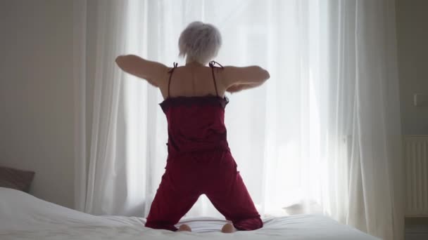 Wide shot back view slim mature woman stretching sitting on bed in the morning. Happy rested Caucasian lady waking up in bedroom indoors at home. Resting and lifestyle. — Stock Video