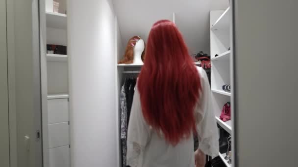En ung transsexuell man i röd peruk går in i garderoben och väljer kläder i slow motion. Självsäker vit icke-binär HBTQ-person som väljer kläder inomhus hemma. Mode och individualitet. — Stockvideo