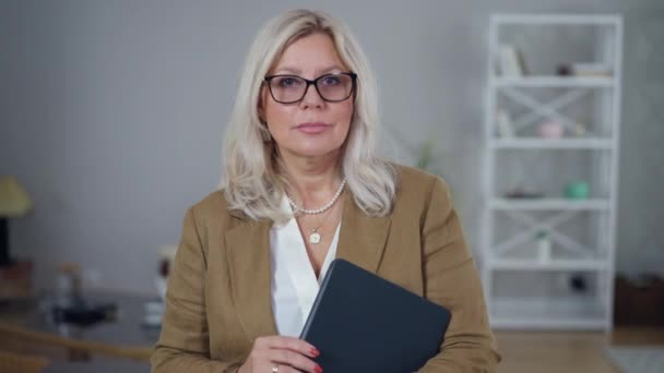 Caméra en direct zoom arrière en tant que psychologue féminine grave avec tablette debout au bureau à la table. attrayant mi-adulte caucasien intelligent femme dans des lunettes posant à l'intérieur. — Video