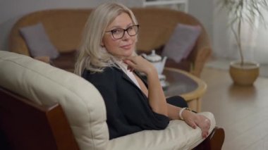 Uzman beyaz kadın psikolog ofisteki rahat koltukta oturmuş kameraya gülümsüyor. İçeride poz veren, kendine güvenen, gözlüklü profesyonel kadın portresi..