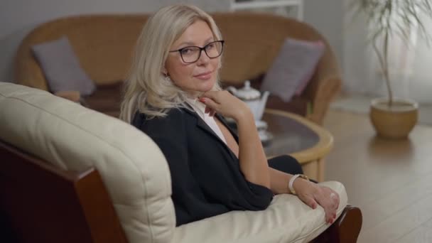 Expert běloška psycholožka sedí na pohodlném křesle v kanceláři a dívá se na usmívající se kameru. Portrét sebevědomé profesionální ženy v brýlích pózující uvnitř. — Stock video