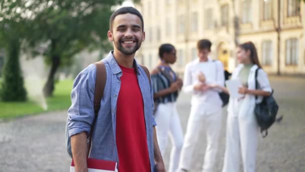 Smiling tampan Timur Tengah mahasiswa muda berpose di kampus dengan kabur multietnis groupmates berbicara di latar belakang. potret pria bahagia yang percaya diri berdiri di luar melihat kamera — Stok Video