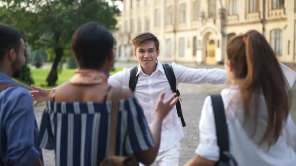 Joyeux jeune homme caucasien courant au ralenti vers un groupe d'amis multiethniques embrassant des camarades de groupe. Beau étudiant excité de rencontrer des amis sur le campus universitaire. Éducation et mode de vie. — Video