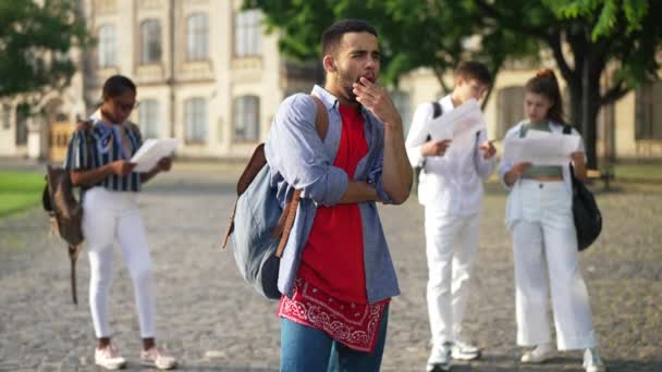Τεμπέλης βαρεθεί Μέση Ανατολή φοιτητής στέκεται σε εξωτερικούς χώρους πανεπιστημιούπολη με σπασίκλες groupmates μάθηση στο παρασκήνιο. Πορτρέτο του αδιάφορου νεαρού που χασμουριέται σε αργή κίνηση. Εκπαιδευτική τεμπελιά. — Αρχείο Βίντεο