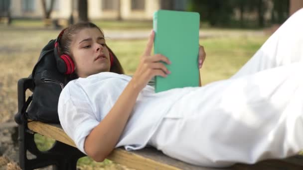 Uvolněná pozitivní mladá žena ležící na lavičce na kolejích a poslouchající hudbu ve sluchátkách. Portrét sebevědomé krásné štíhlé běloška studenta těší koníček na přestávce venku. Zpomalený pohyb. — Stock video