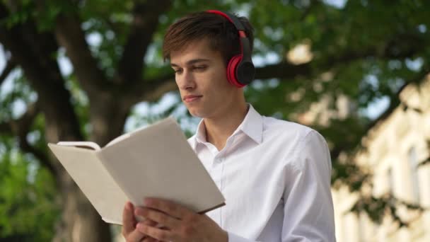 Portret przystojnego brunetka młodego człowieka czytającego książkę na pamięć, uczącego się na świeżym powietrzu w słoneczny dzień. Skoncentrowany student uniwersytetu w słuchawkach uczący się na podwórku. Zwolniony ruch. — Wideo stockowe