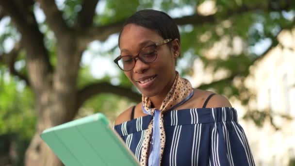 Gözlüklü, tabletli, dışarıda kameraya gülümseyen kendinden emin Afro-Amerikan bayan öğrenci. Güneşli bir günde üniversite bahçesinde poz veren kendinden emin zeki bir kadının portresi. Yavaş çekim. — Stok video