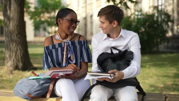 Happy young interracial couple of students sitting on bench outdoors turning looking at camera waving in slow motion. Portrait of confident African American woman and Caucasian man talking at college. — Stock Video