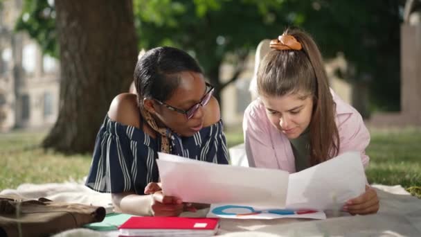 Positive Caucasian and African American young beautiful women lying on sunny university lawn talking in slow motion. Portrait of confident students friends analyzing paperwork discussing lesson. — Stock Video