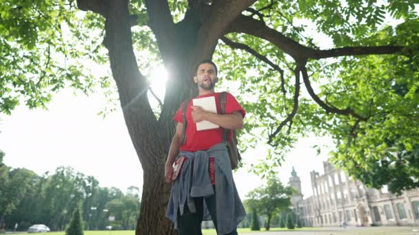 Junger Student aus dem Nahen Osten steht gähnend und streckend im Sonnenlicht an einem Baum auf dem Universitätshof. Porträt eines gutaussehenden bärtigen Mannes, der an einem sonnigen Morgen im Freien wartet. Zeitlupe. — Stockvideo