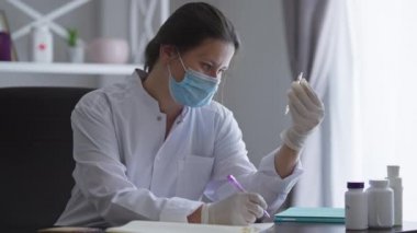 Covid-19 yüz maskeli profesyonel kadın bilim adamı uyuşturucu ampulleriyle masada oturup kalemle yazı yazıyor. İlacın yan etkilerini ve verimliliğini analiz eden beyaz bir kadın..