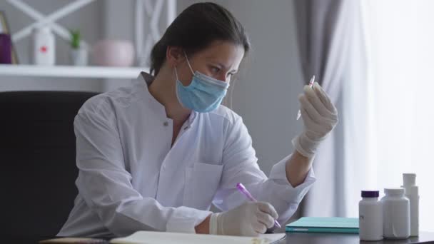 Covid-19 yüz maskeli profesyonel kadın bilim adamı uyuşturucu ampulleriyle masada oturup kalemle yazı yazıyor. İlacın yan etkilerini ve verimliliğini analiz eden beyaz bir kadın.. — Stok video