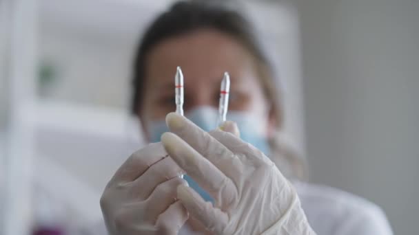 Detailní ampulky s tekutými drogami v ženských rukou v rukavicích s rozmazanou běloškou v Covid-19 obličejové masce na pozadí. Mladý lékař vědec zkoumá léky uvnitř v nemocnici. — Stock video