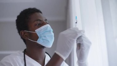 Coronavirus yüz maskesi takan yakın plan Afrika kökenli Amerikalı doktor elinde Covid-19 aşısı olan bir şırınga tutuyor. Hastahanede salgın aşısı olan ciddi bir adam. Vax konsepti.